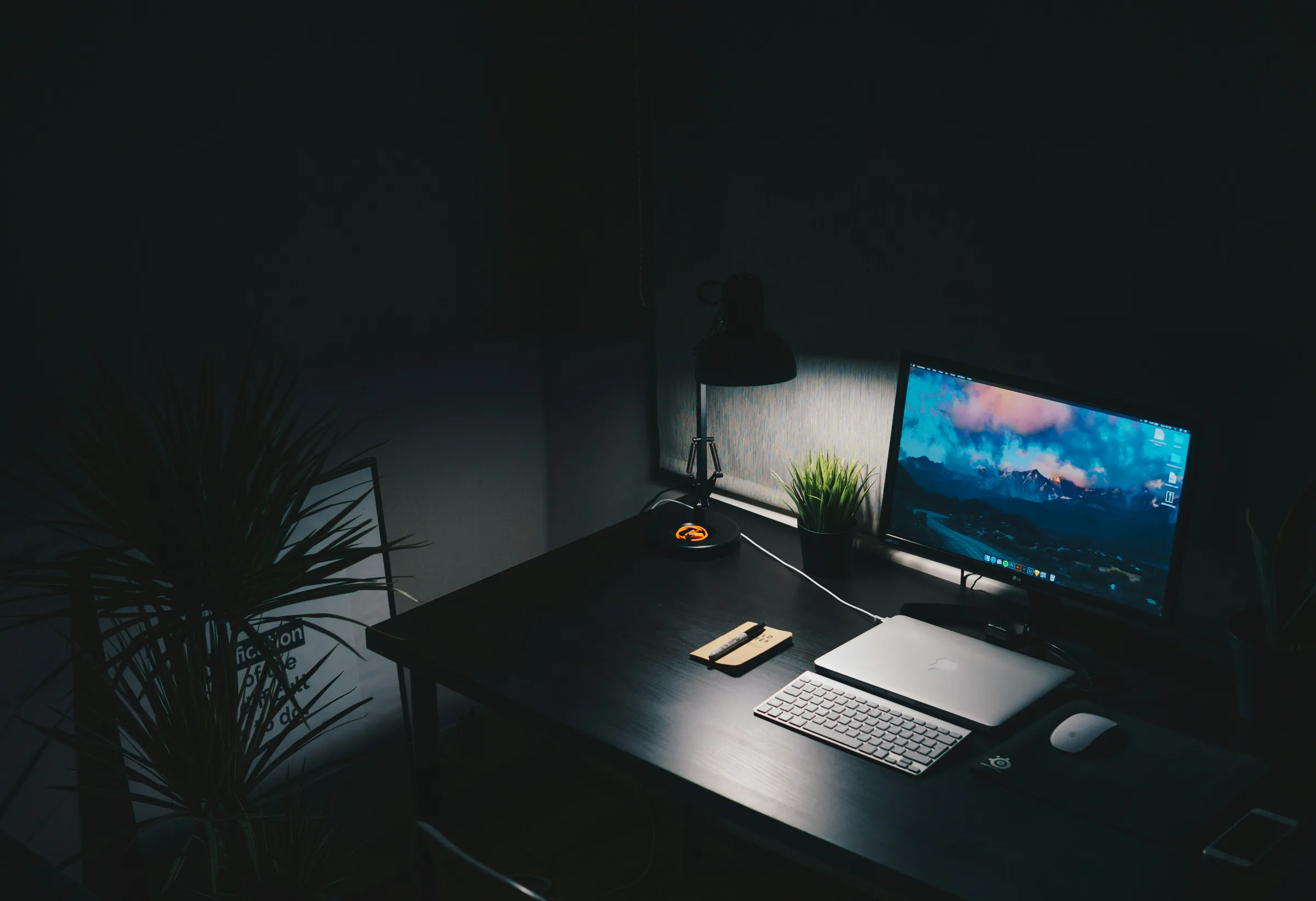A desk in a dark room under one lamp: a monitor, a closed laptop, a keyboard and a small plant